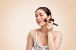 Young brunette woman applies makeup to her cheek to try to conceal rosacea
